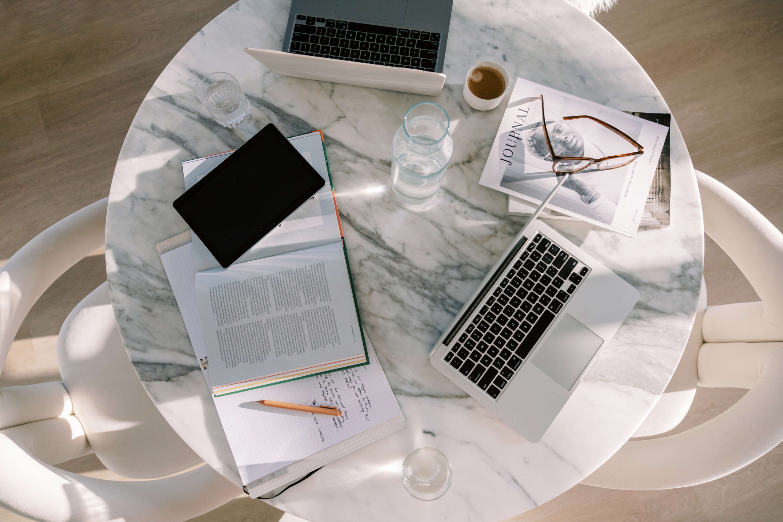 Top-down view of a workspace with laptops, notebooks, glasses, and coffee on a marble table.