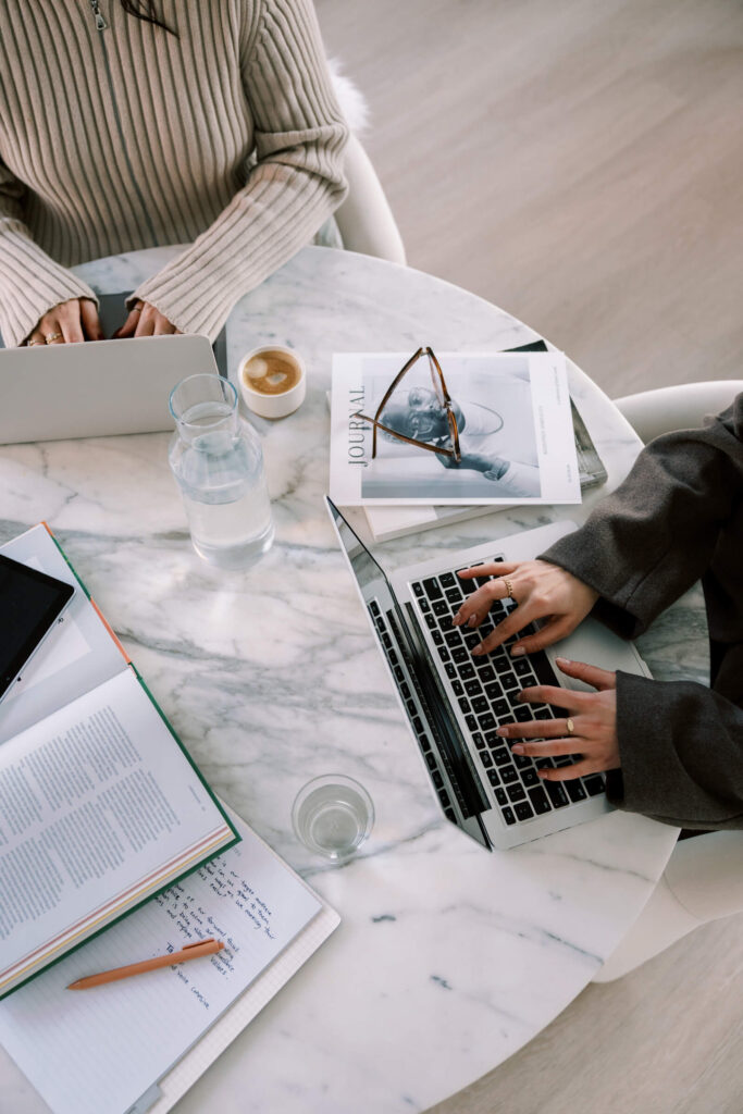 Close-up of hands typing on laptops with journals, coffee, and water on a modern workspace.